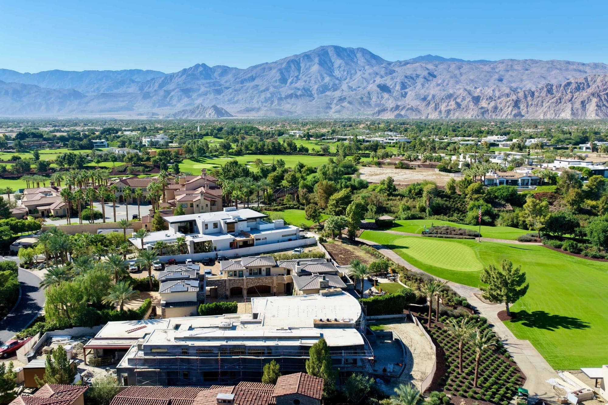 An aerial view of a low white desert estate in La Quinta along a green fairway, with the Santa Rosa mountains behind it.