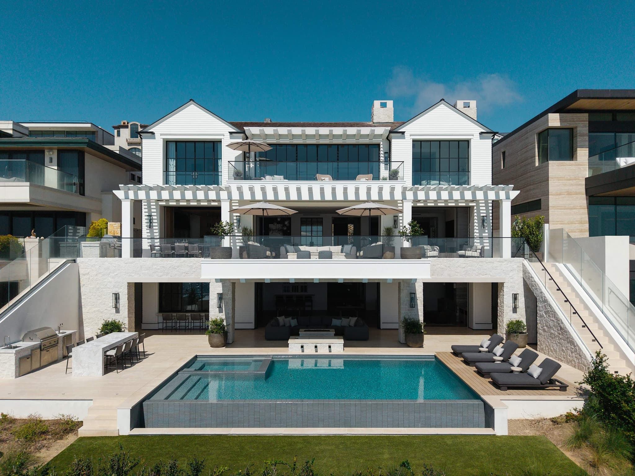 A white stone and shingle oceanfront home at The Strand in Dana Point, its pergola-shaded terraces and pool stepping down to the sand beneath walls of glass.