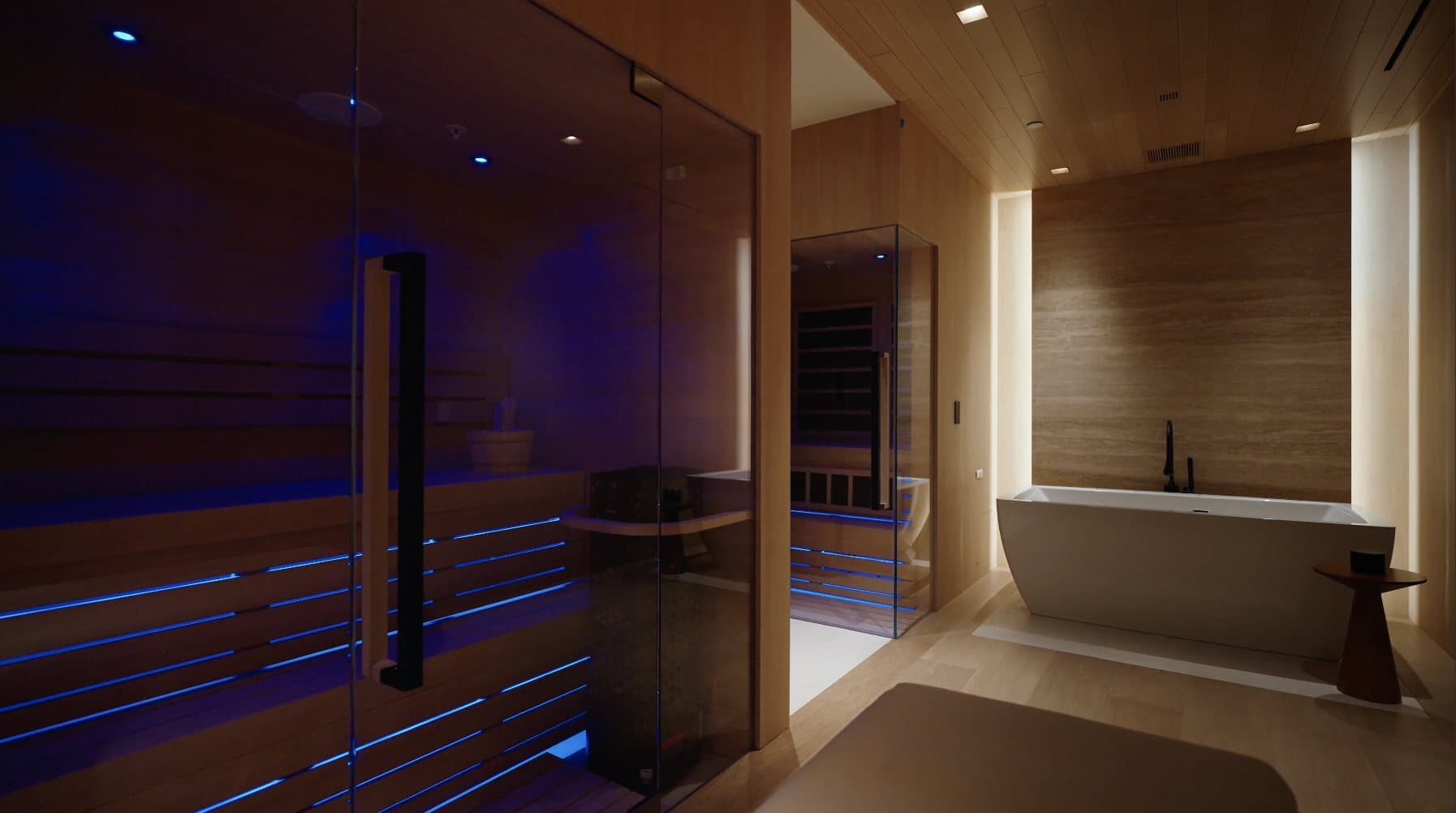 A wellness suite in a Genova home: a glass-fronted sauna glowing soft blue beside a travertine wall and a freestanding soaking tub.